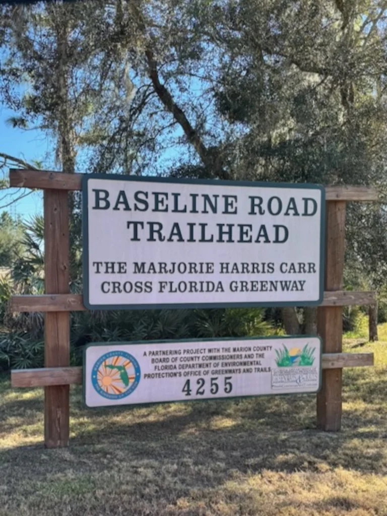 The Baseline Road Trailhead sign reading 'The Marjorie Harris Carr Cross Florida Greenway' with trees and palmetto in the background.